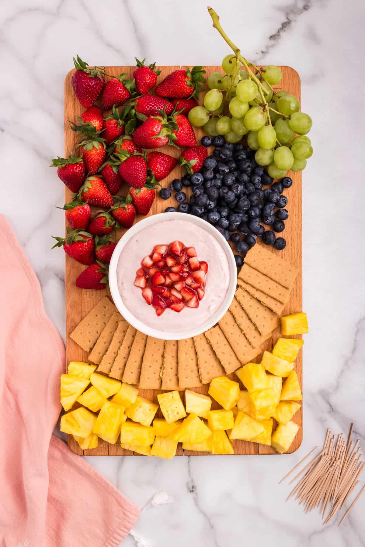 A wooden board beautifully arranged with strawberries, blueberries, green grapes, pineapple chunks, and graham crackers surrounding a bowl of creamy strawberry dip topped with a glossy strawberry mixture.