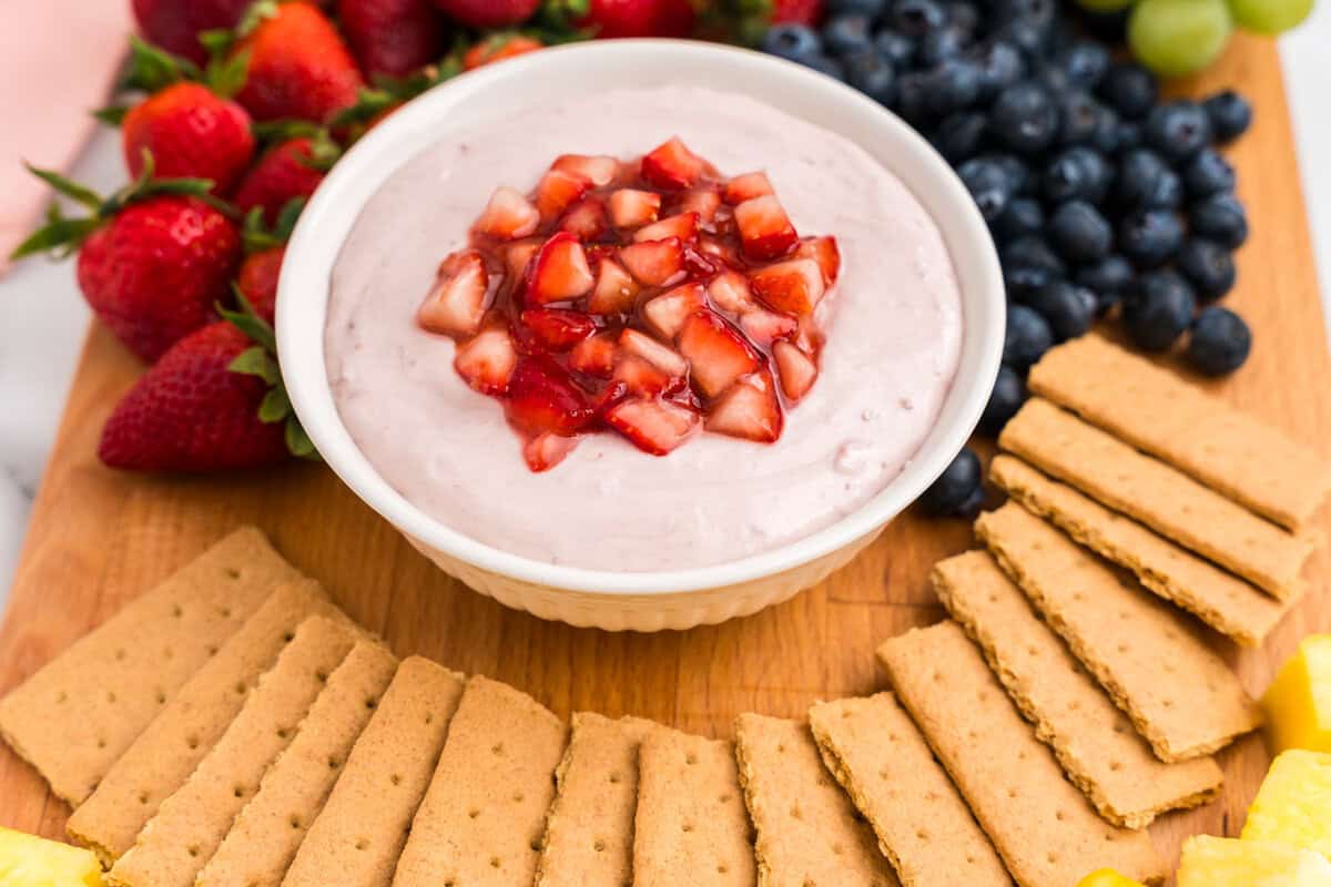 A close-up of the completed serving board featuring a white bowl filled with pink strawberry dip topped with chopped strawberries, surrounded by fresh fruits and graham crackers.