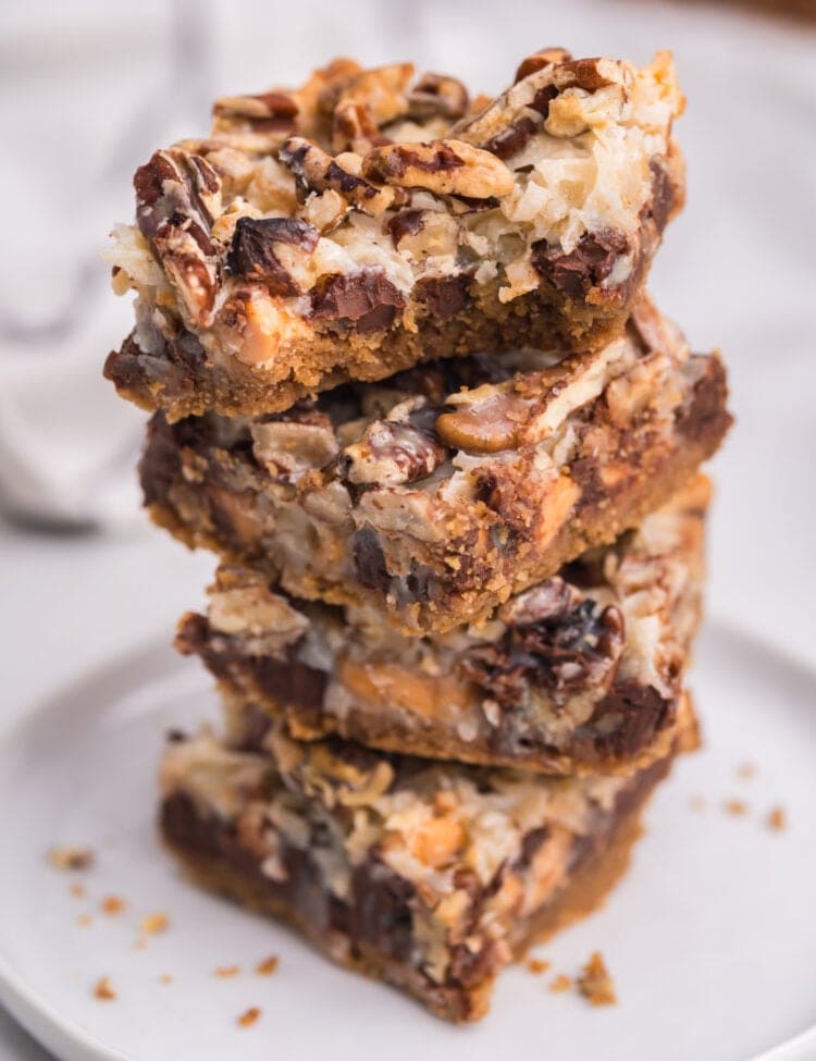 A close-up of a stack of cut 7-layer bars on a white plate, showcasing distinct layers of graham cracker crust, melted chips, coconut, and toasted pecans.