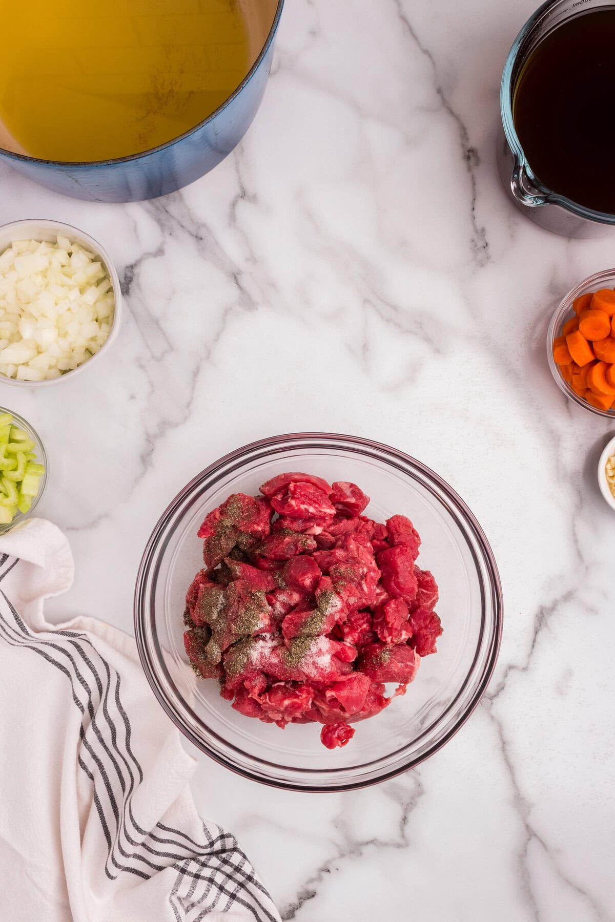 A glass bowl filled with raw beef chunks seasoned with salt and pepper sits on a marble countertop. Surrounding it are small bowls of diced onions, carrots, celery, and garlic. A large pot with oil and a measuring cup filled with beef broth are also visible.