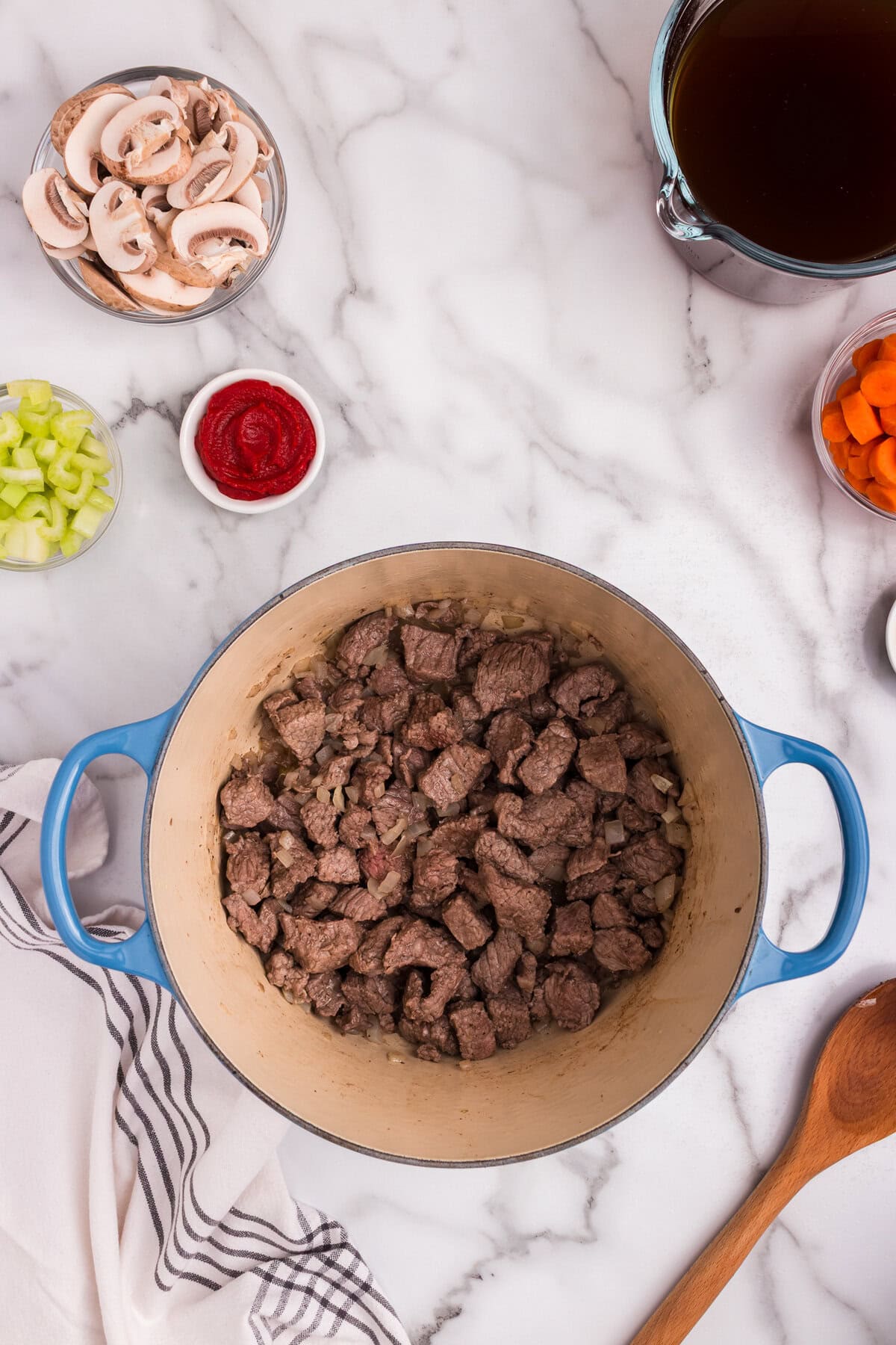 Browned beef chunks are sautéing in a blue Dutch oven on a marble surface. The pot is surrounded by ingredients including sliced mushrooms, tomato paste, celery, carrots, and beef broth.