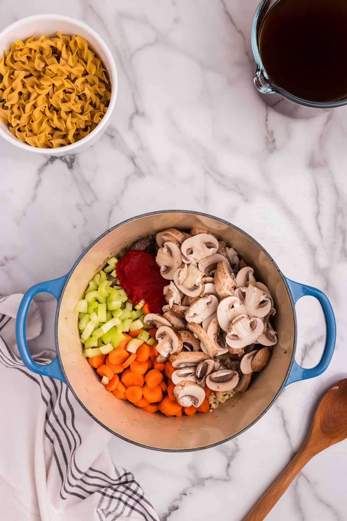A Dutch oven filled with browned beef now topped with sliced mushrooms, diced carrots, chopped celery, minced garlic, and tomato paste. Ingredients are added but not yet stirred together.