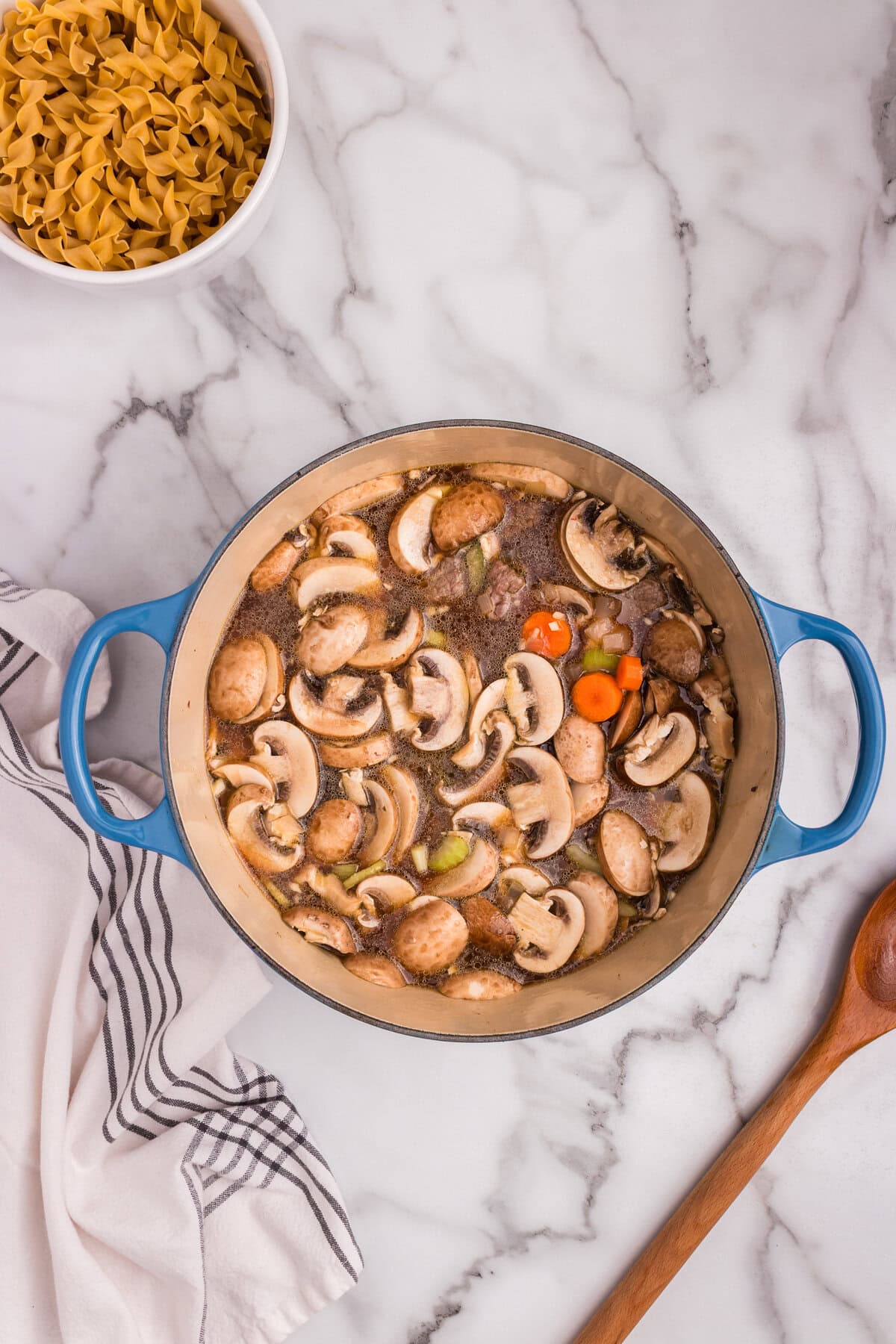 The same Dutch oven is now filled with beef broth, partially submerging mushrooms, carrots, and other vegetables. Ingredients are beginning to simmer and cook together.
