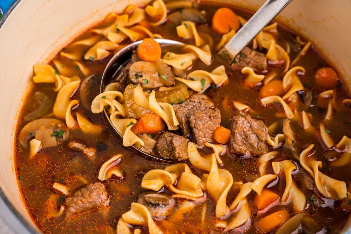 A ladle scoops a hearty portion of beef noodle soup from a large Dutch oven. The scoop includes beef chunks, egg noodles, carrots, celery, mushrooms, and is garnished with fresh herbs.