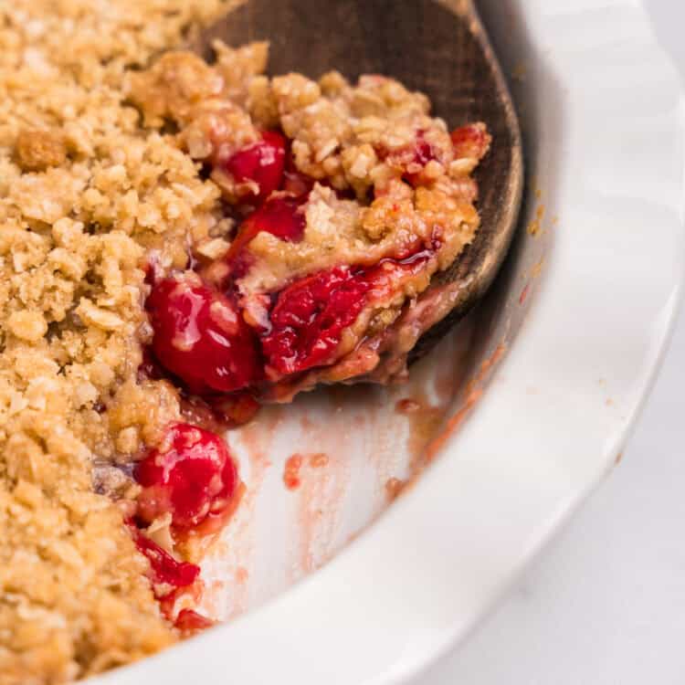 A wooden spoon lifting a spoonful of cherry crumble from the dish, showing the texture and filling.