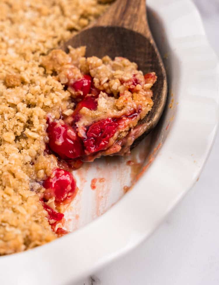 A wooden spoon lifting a spoonful of cherry crumble from the dish, showing the texture and filling.