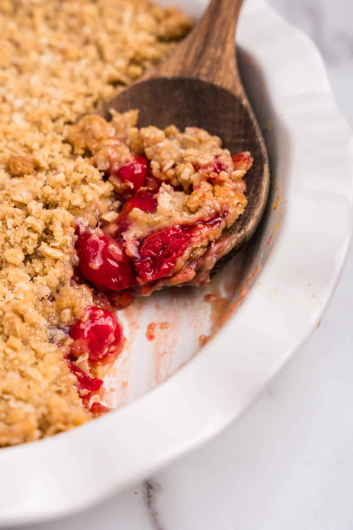 A wooden spoon lifting a spoonful of cherry crumble from the dish, showing the texture and filling.

