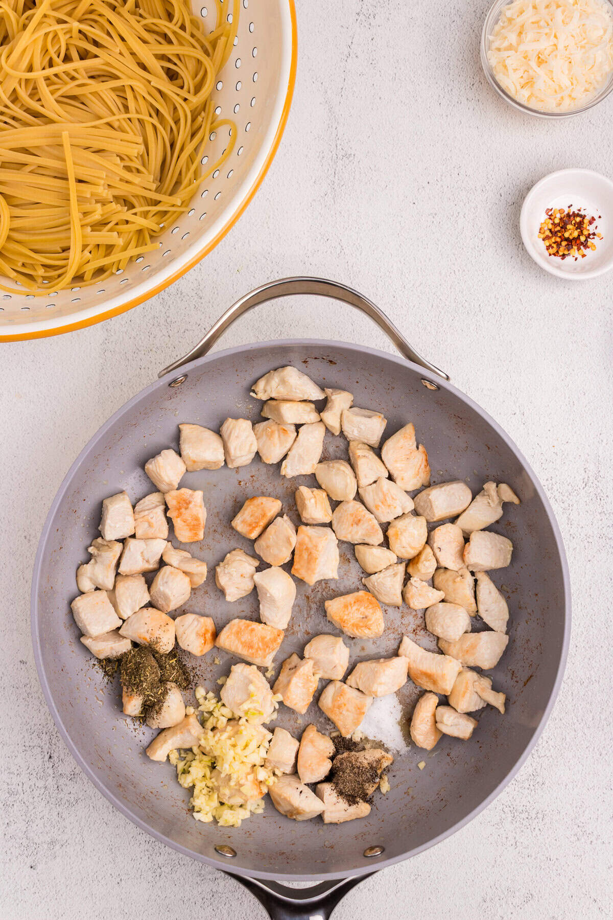 Raw linguine pasta in a colander next to a skillet with browned chicken chunks, minced garlic, herbs, salt, and pepper being added.
