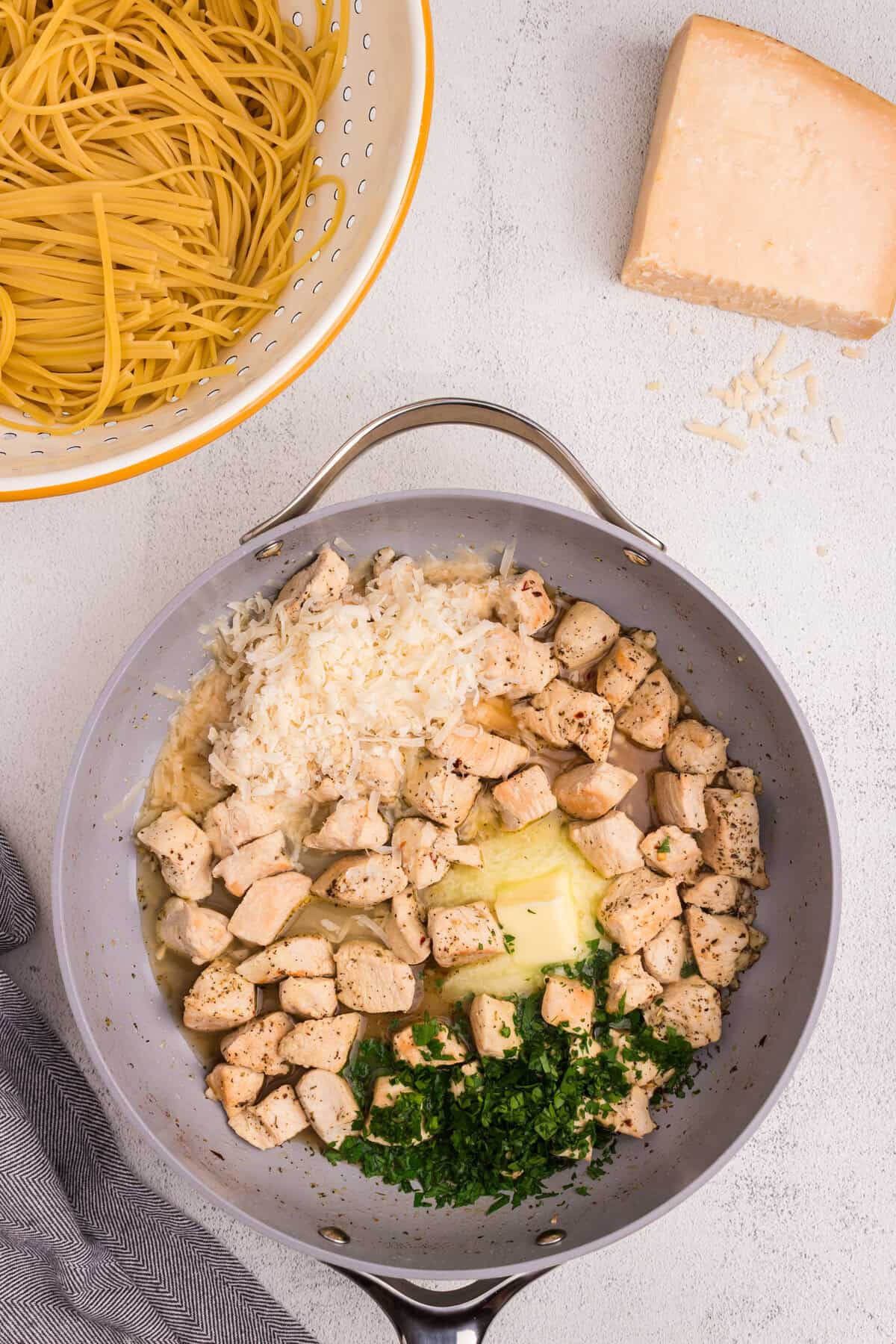 Skillet with cooked chicken, grated cheese, butter, chopped parsley, and broth, next to uncooked pasta and a wedge of Parmesan cheese.