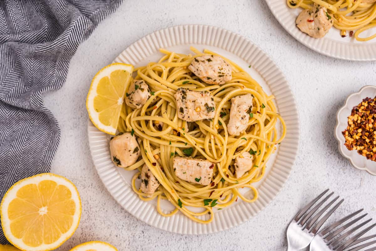 Overhead view of two plates of chicken scampi garnished with lemon slices and red pepper flakes, set beside utensils and a kitchen towel.