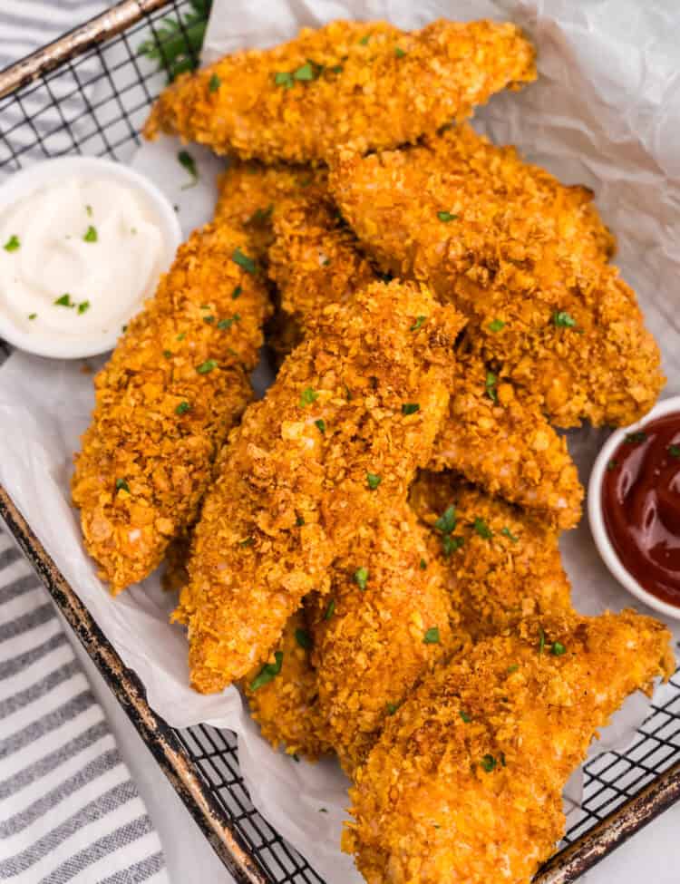 A wire basket lined with parchment paper filled with crispy baked cornflake chicken tenders, garnished with parsley and served with small bowls of ketchup and mayonnaise for dipping.