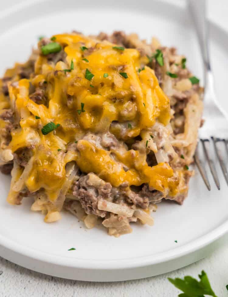 A close-up of a serving of hamburger hashbrown casserole on a white plate with a fork. The dish is topped with melted cheddar cheese and sprinkled with chopped fresh parsley.