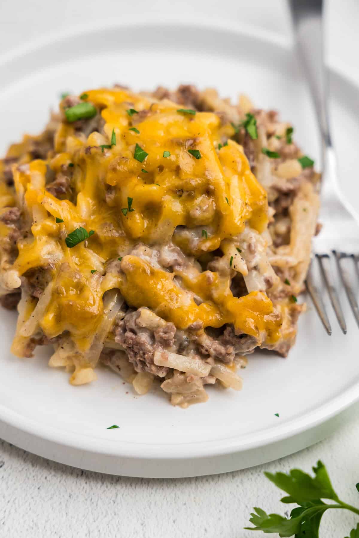 A close-up of a serving of hamburger hashbrown casserole on a white plate with a fork. The dish is topped with melted cheddar cheese and sprinkled with chopped fresh parsley.