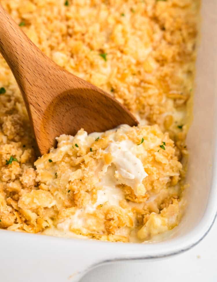 A close-up of a creamy chicken casserole with a golden, crispy cracker topping, being scooped with a wooden spoon from a white baking dish. The casserole appears rich and cheesy, with a crunchy texture on top, garnished with small flecks of green herbs.