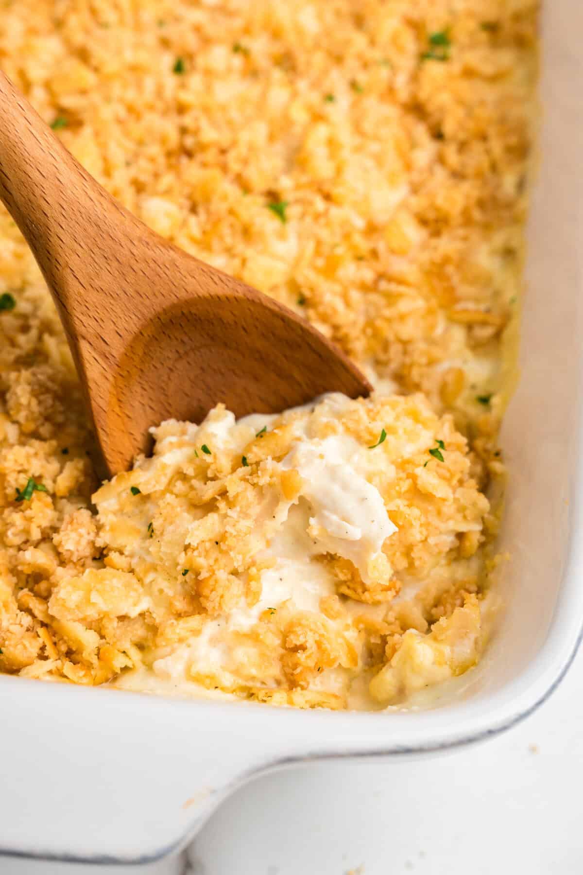 A close-up of a creamy chicken casserole with a golden, crispy cracker topping, being scooped with a wooden spoon from a white baking dish. The casserole appears rich and cheesy, with a crunchy texture on top, garnished with small flecks of green herbs.