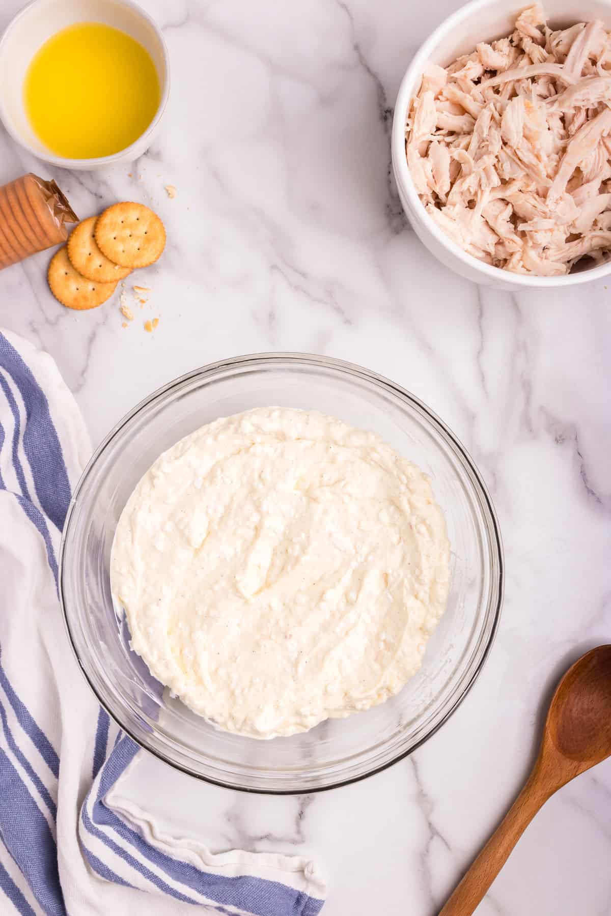 A glass mixing bowl filled with a creamy, blended mixture of ingredients, creating a smooth consistency. Surrounding the bowl are a wooden spoon, a dish of shredded chicken, a small bowl of melted butter, and a few scattered crackers on a marble countertop.
