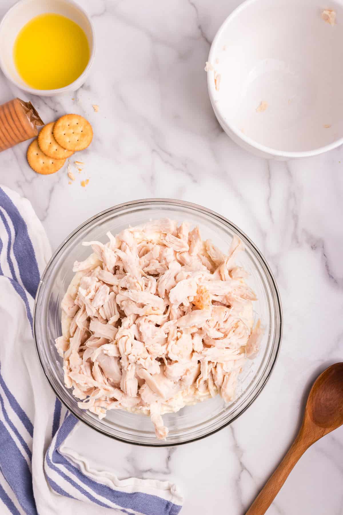 A glass mixing bowl filled with shredded cooked chicken placed on top of a creamy mixture, ready to be combined. Surrounding the bowl are a wooden spoon, an empty white bowl, a small dish of melted butter, and a few scattered crackers on a marble countertop.