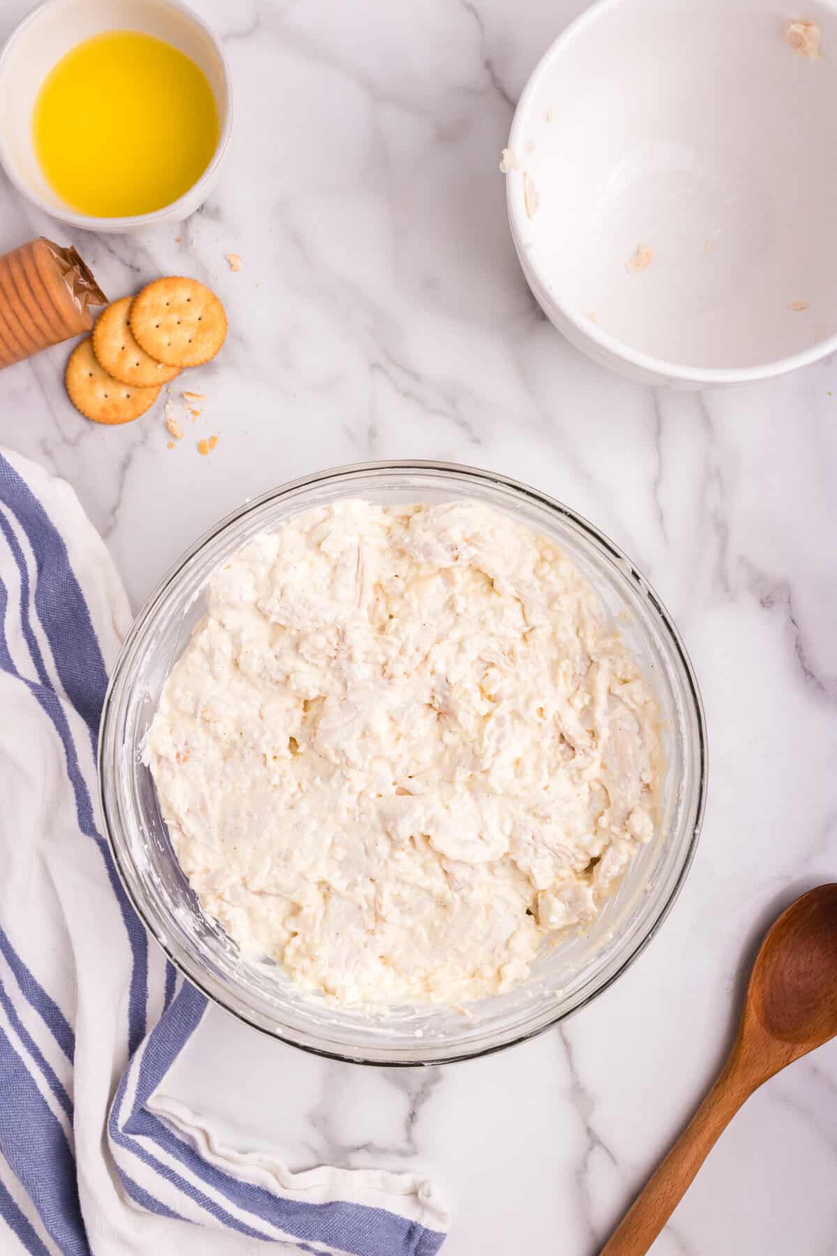 A glass mixing bowl filled with a creamy chicken mixture, fully blended and ready for the next step in the recipe. The bowl is placed on a marble countertop with a wooden spoon, an empty white bowl, a small dish of melted butter, and scattered crackers nearby.