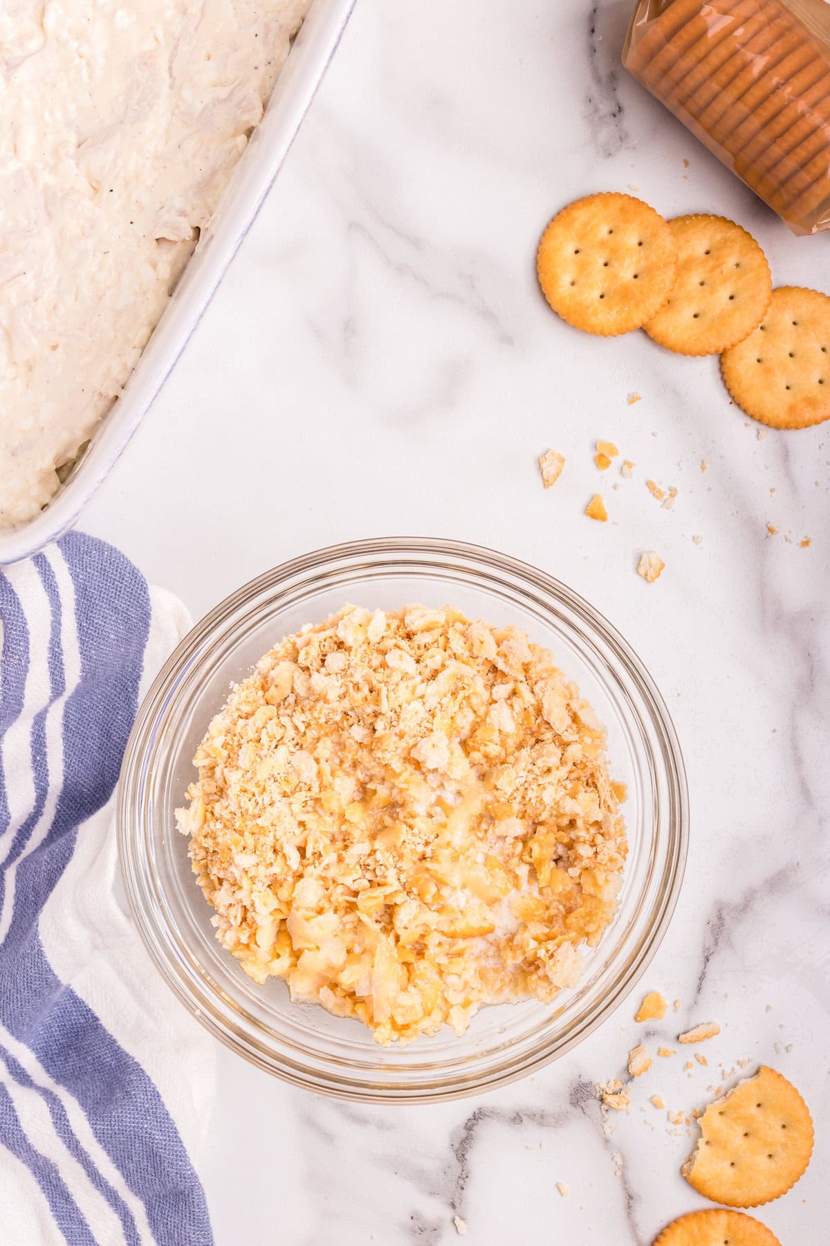 A glass bowl filled with a mixture of crushed buttery crackers and melted butter, ready to be used as a crunchy topping for the casserole. Nearby, a partially visible baking dish contains the creamy chicken mixture, with a sleeve of crackers and a few broken pieces scattered on a marble countertop.