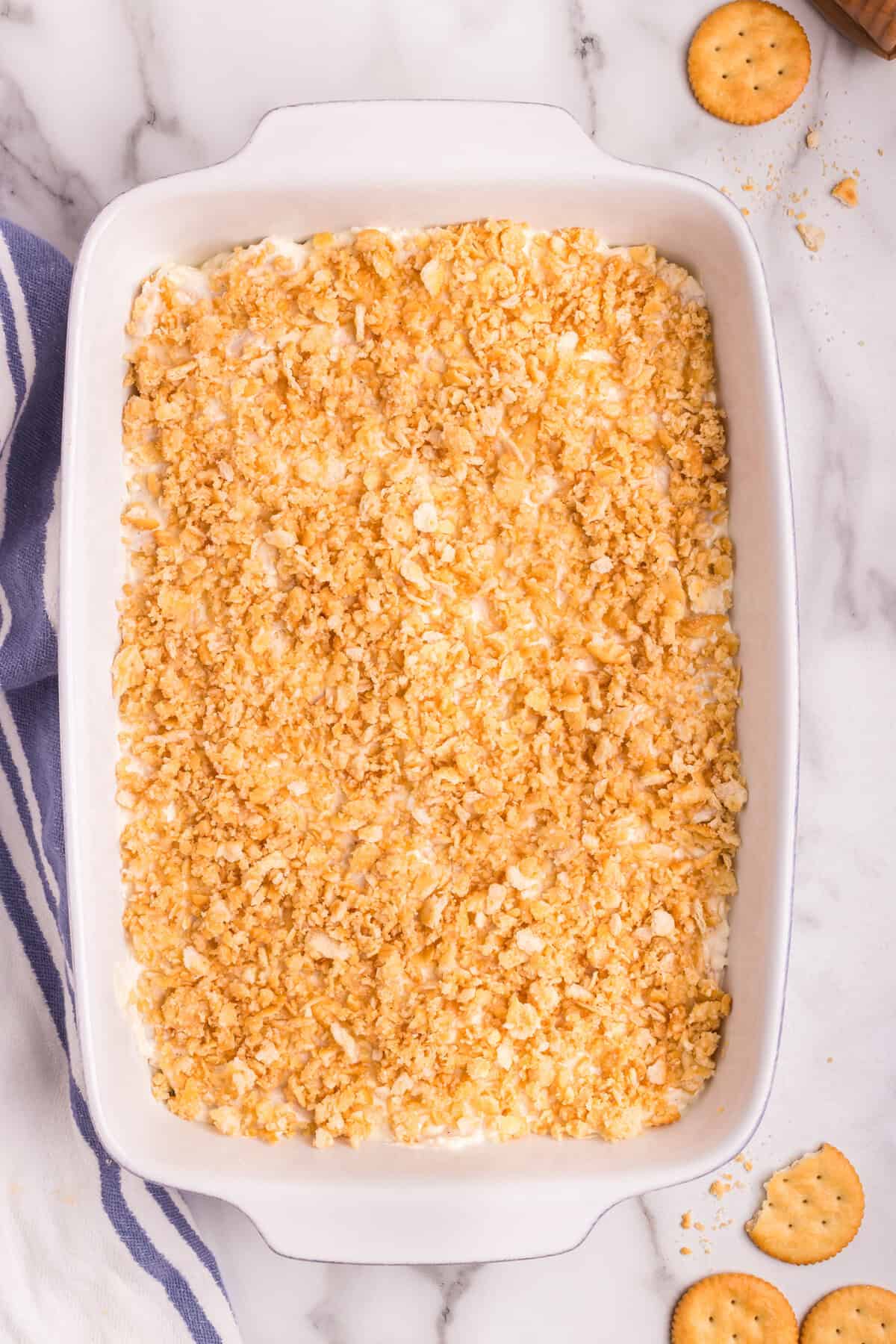 A white baking dish filled with unbaked Million Dollar Chicken Casserole, topped with a golden, buttery crushed cracker mixture. The dish is placed on a marble countertop with a few whole and broken crackers scattered nearby.