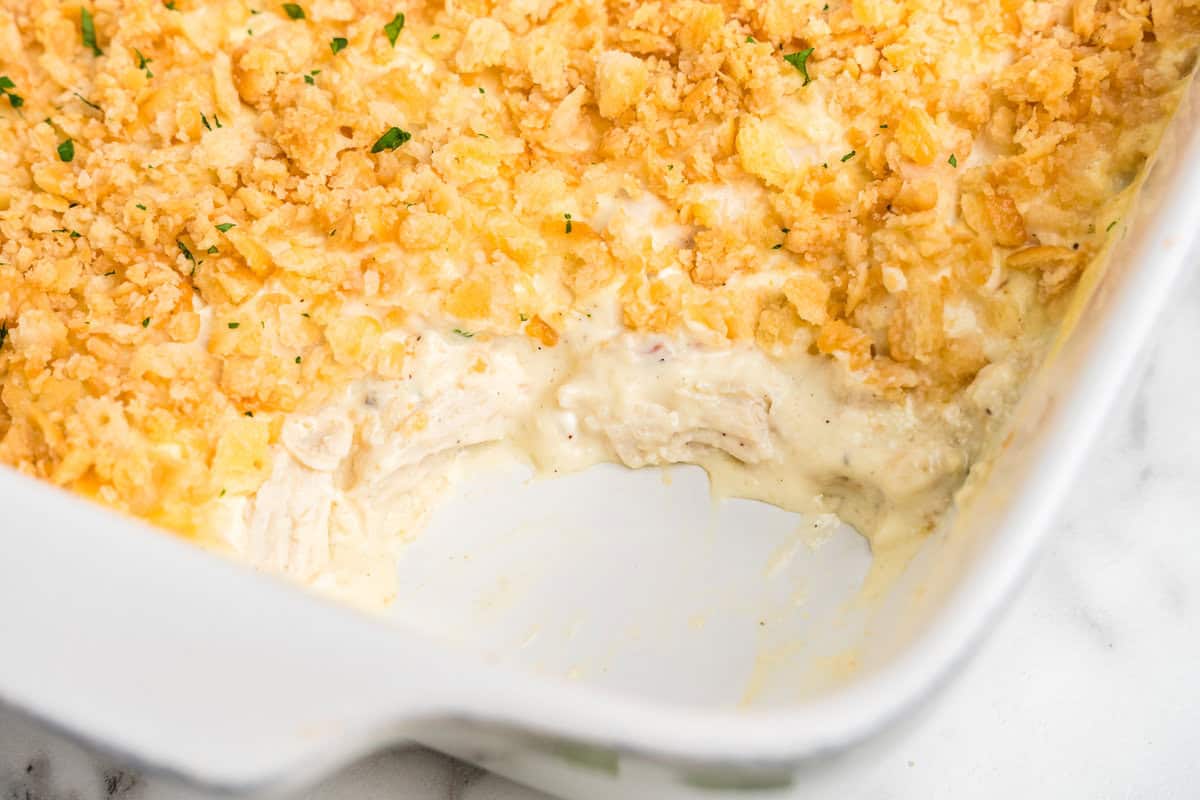 A partially eaten chicken casserole in a white baking dish, showing its creamy, cheesy interior with shredded chicken and a golden, crispy cracker topping. Small flecks of green herbs are sprinkled on top for garnish.
