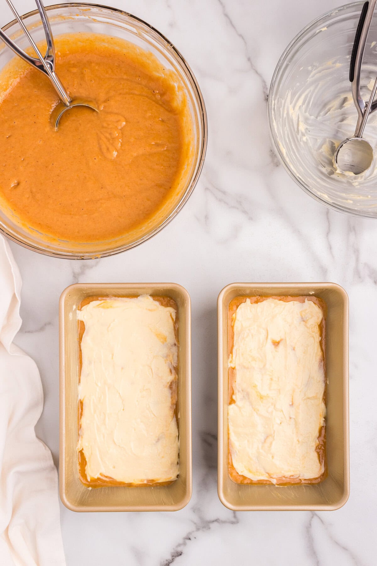 Loaf pans with cream cheese filling spread smoothly over the first layer of pumpkin batter, with bowls of batter and a cookie scoop beside them.