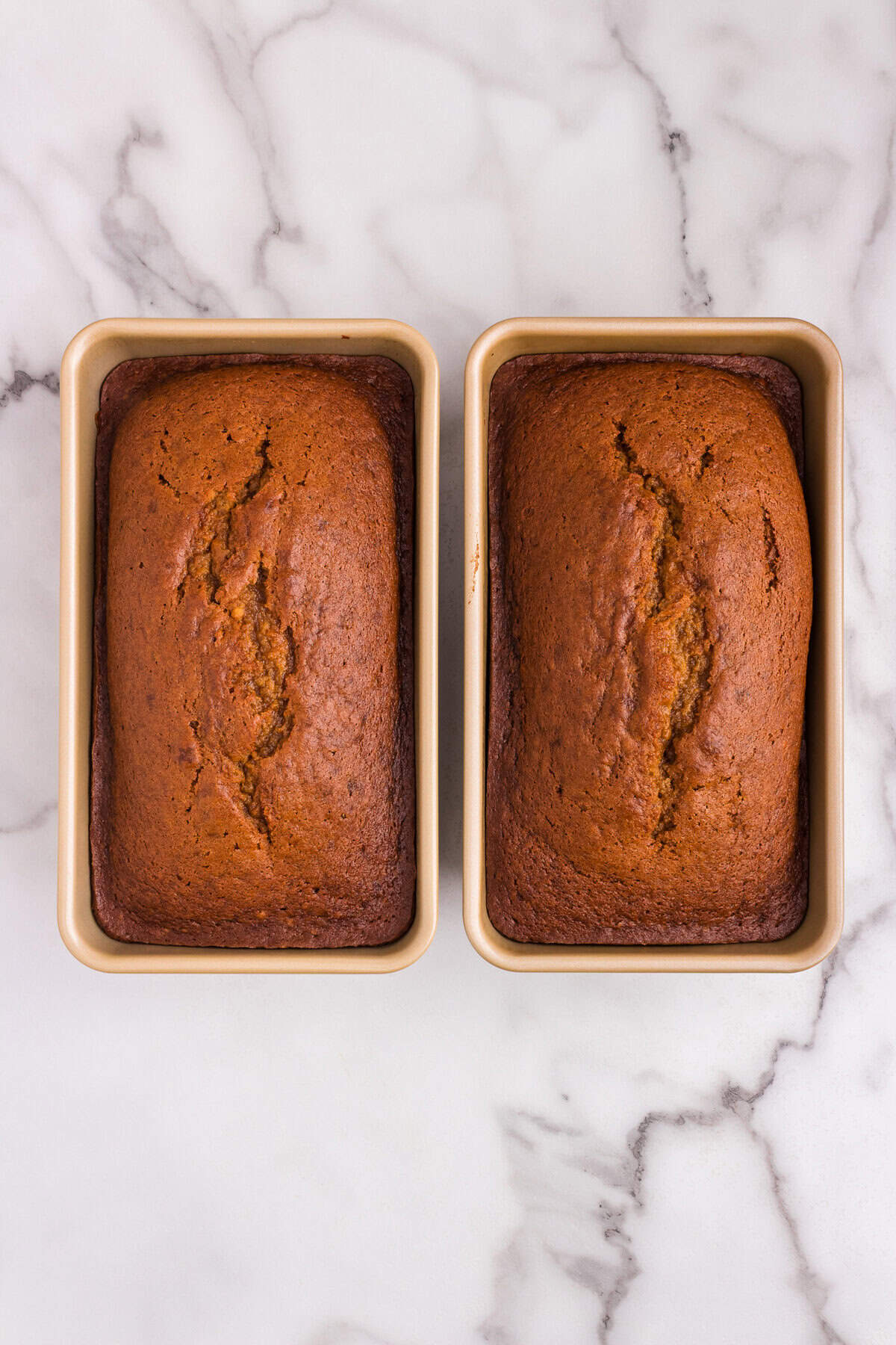 Two freshly baked loaves of pumpkin bread in pans, golden brown and cracked slightly on top, resting on a marble countertop.