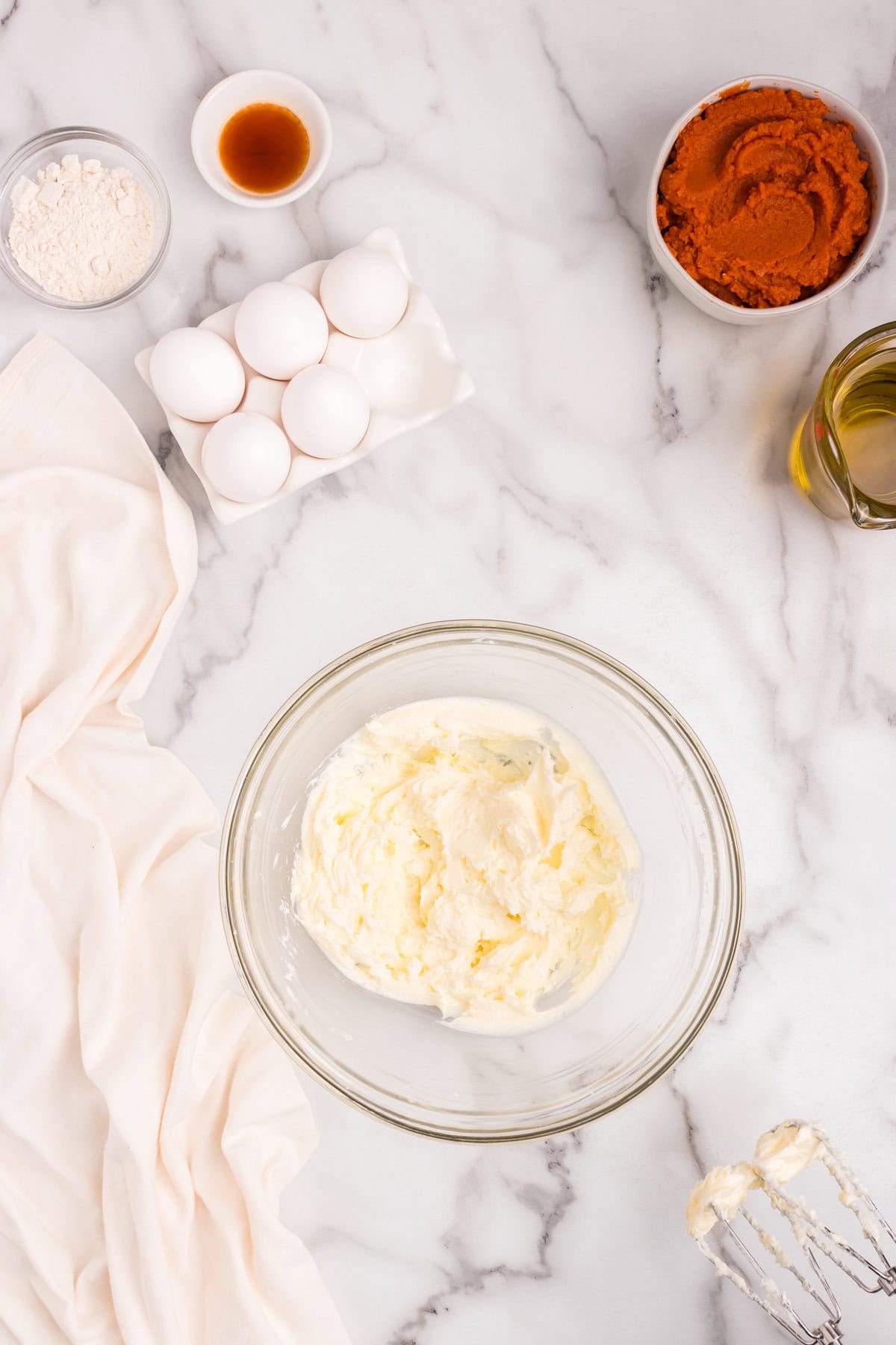 Glass mixing bowl with softened cream cheese being prepared; eggs, vanilla, and flour are nearby on a marble surface.