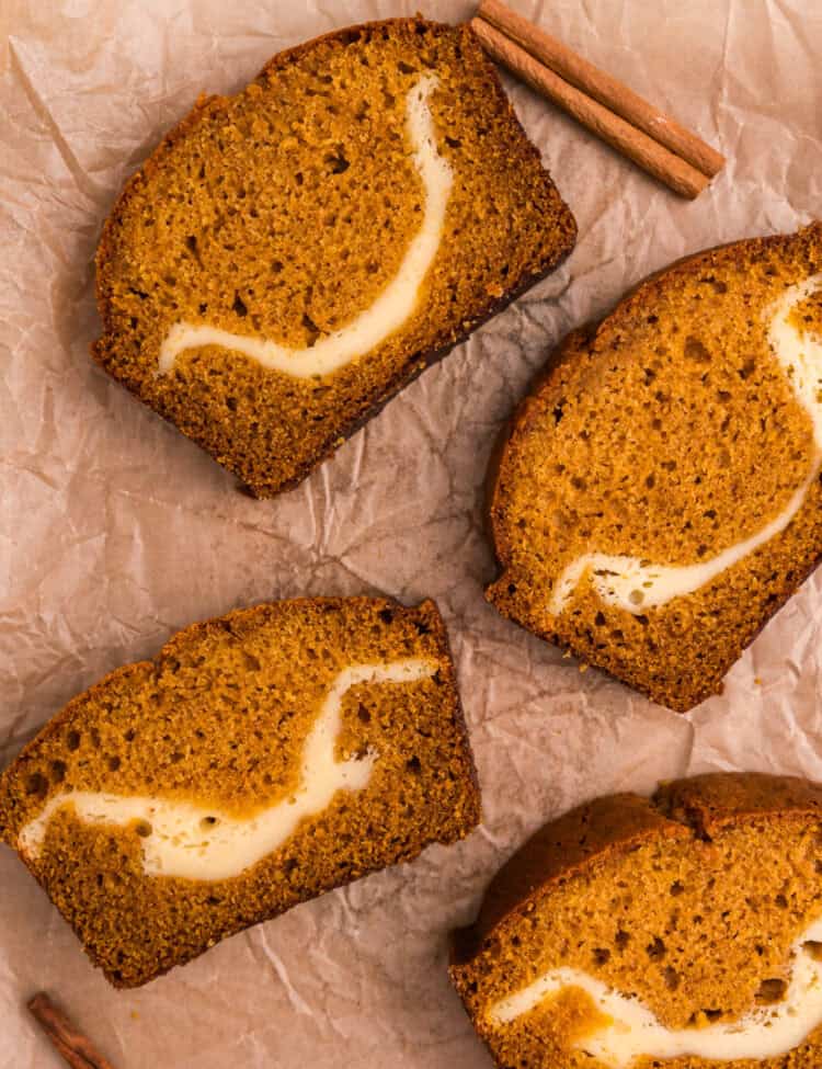 Four slices of pumpkin cream cheese bread arranged on parchment paper with cinnamon sticks, showing the marbled cream cheese filling in each slice.