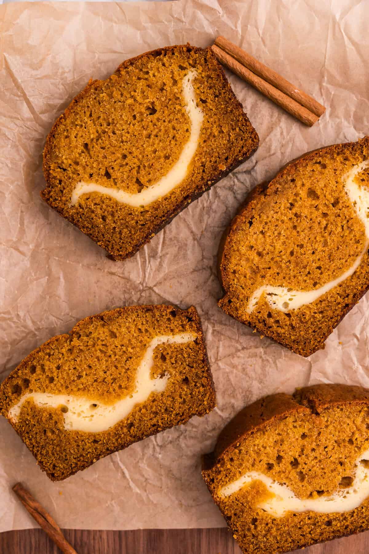 Four slices of pumpkin cream cheese bread arranged on parchment paper with cinnamon sticks, showing the marbled cream cheese filling in each slice.