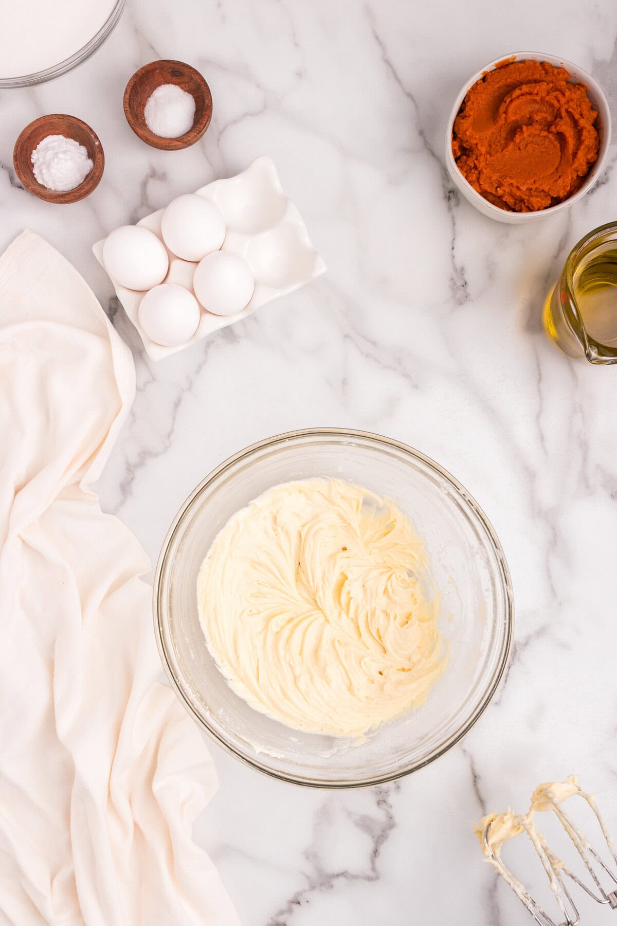 Cream cheese mixture fully blended to a smooth consistency in a glass bowl on a marble countertop with baking ingredients surrounding it.