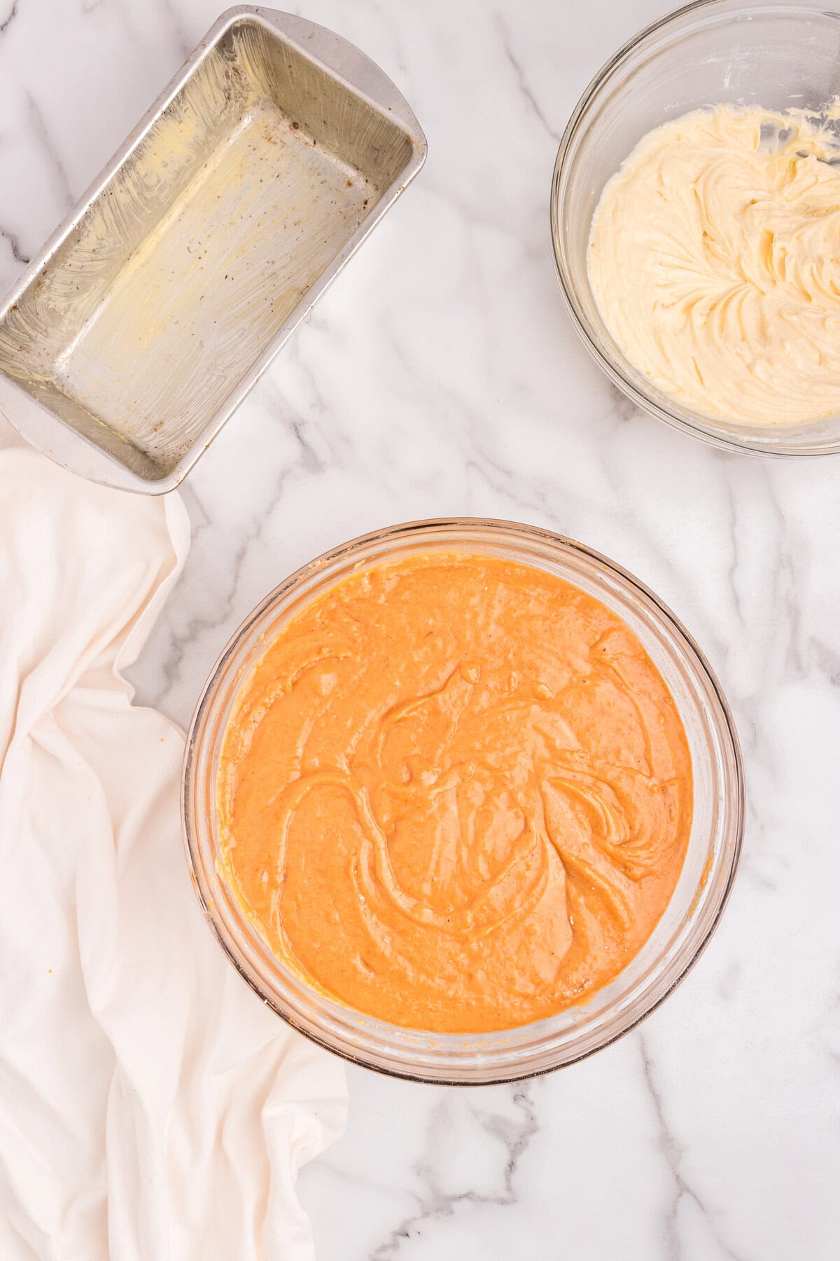 Top-down view of a greased loaf pan next to bowls of pumpkin batter and cream cheese filling on a marble surface.