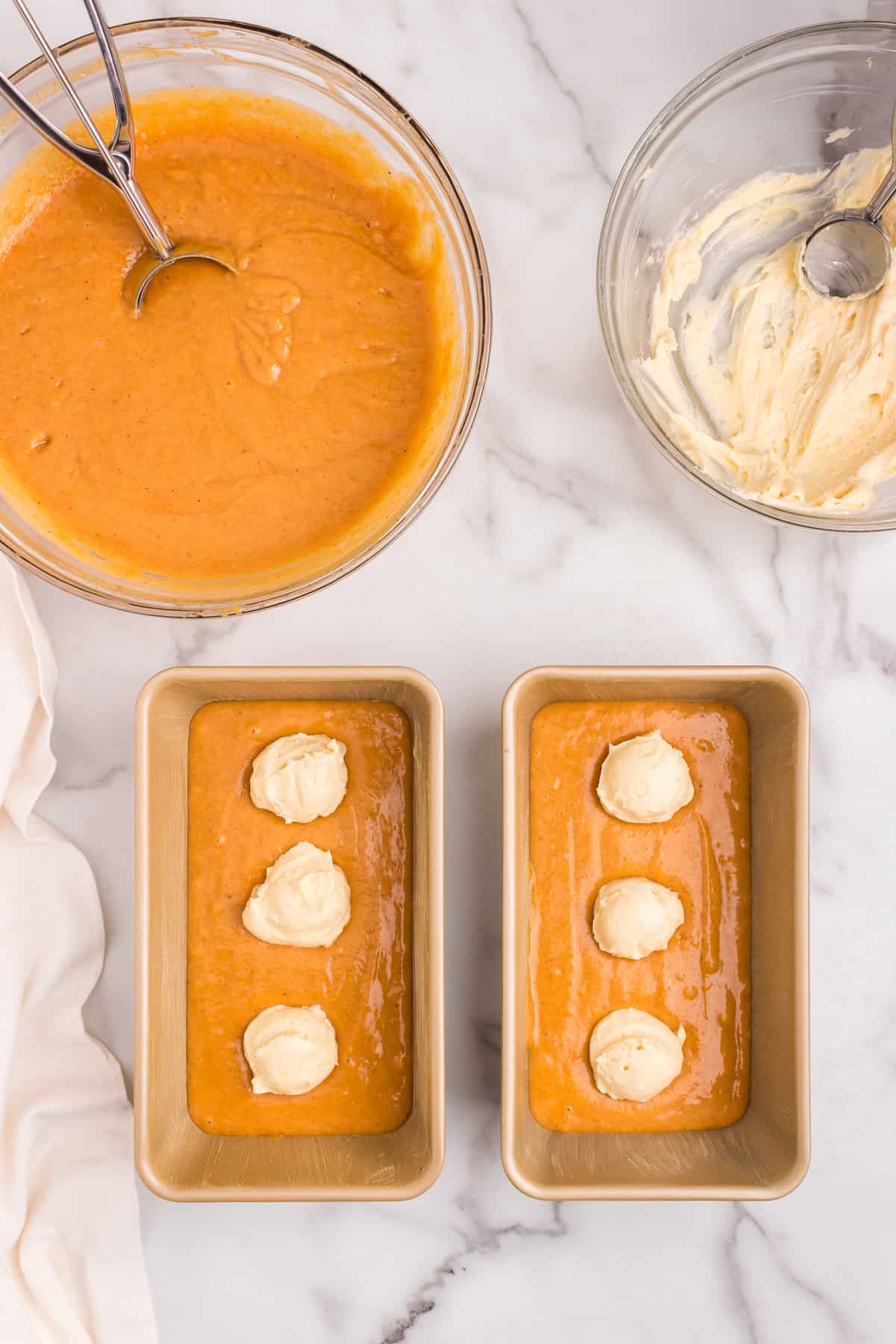 Two loaf pans partially filled with pumpkin batter, each with evenly spaced dollops of cream cheese filling on top. Mixing bowls with remaining batters in background.
