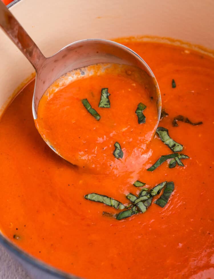 Close-up of a ladle serving smooth tomato soup from the Dutch oven, topped with strips of fresh basil.