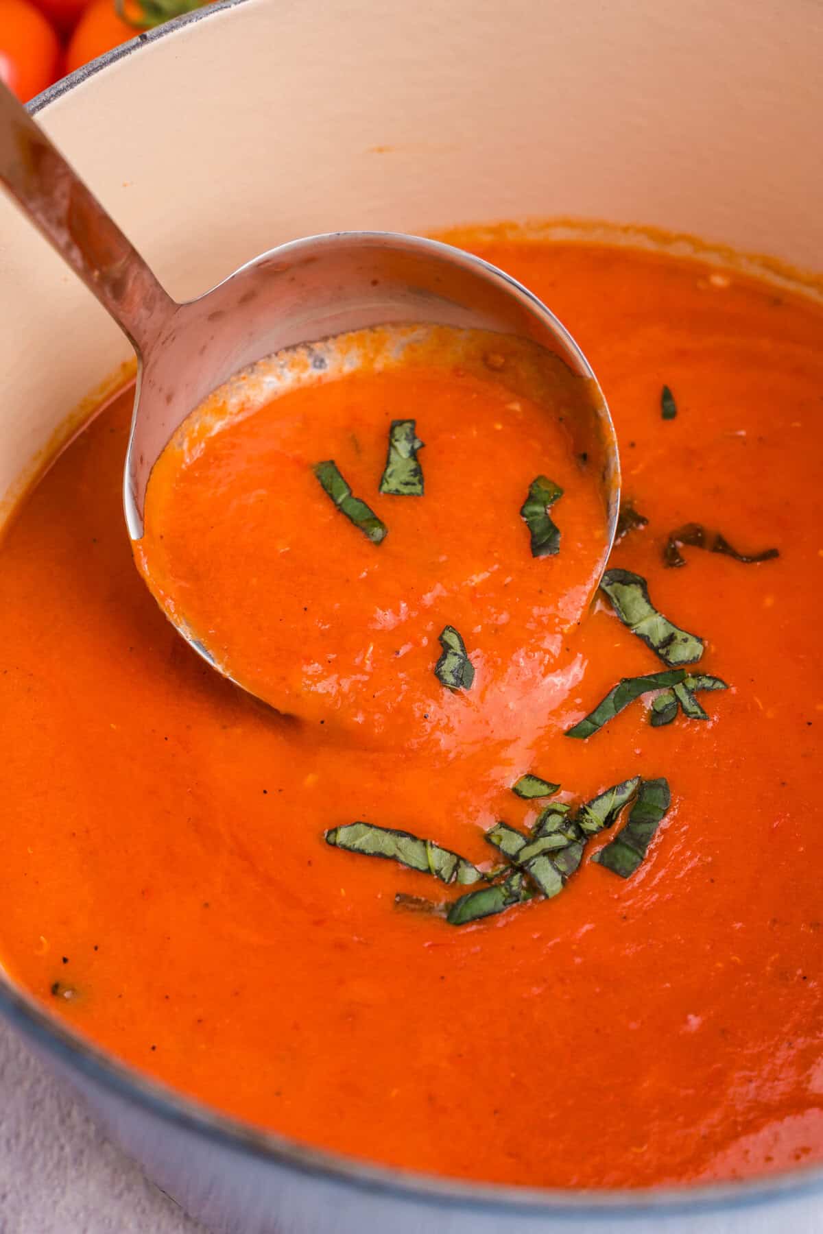  Close-up of a ladle serving smooth tomato soup from the Dutch oven, topped with strips of fresh basil.


