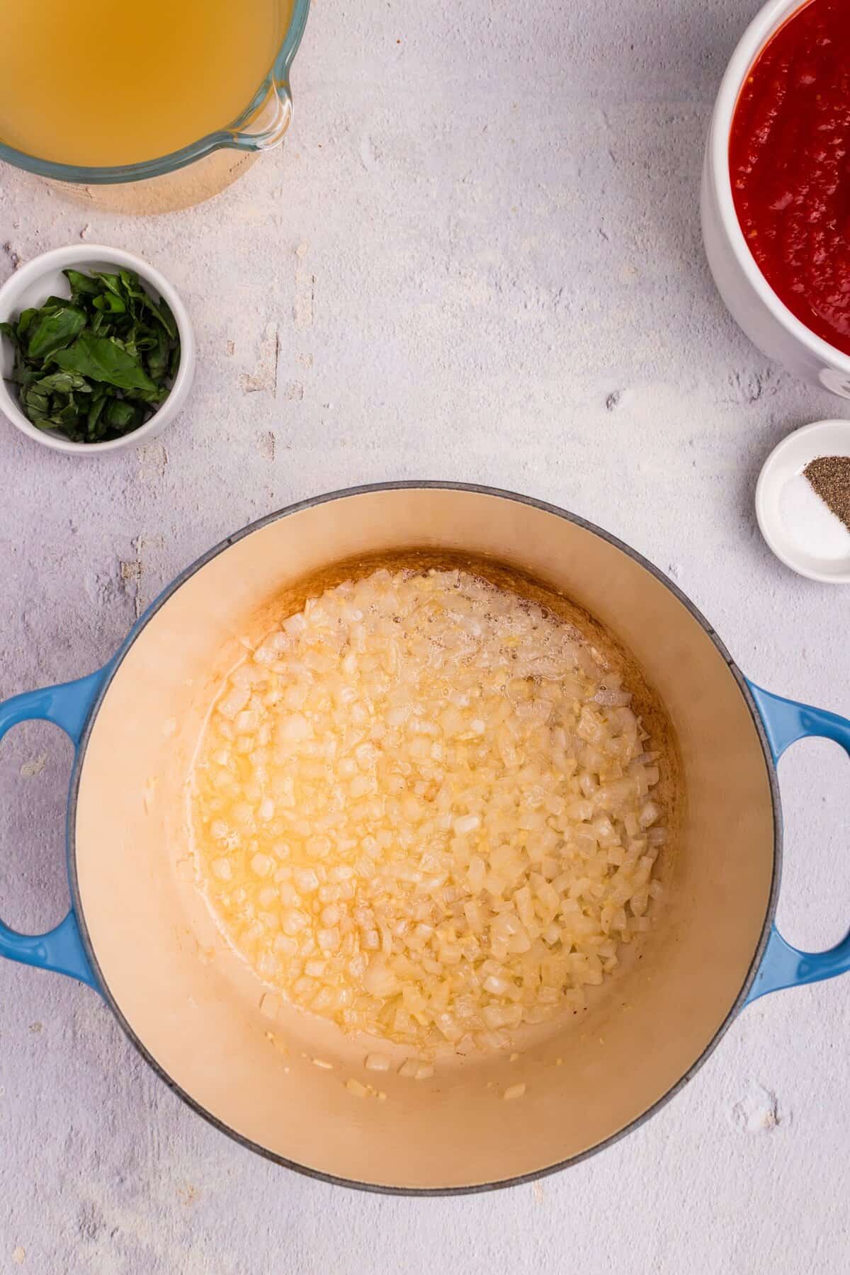 Diced onions sautéing in butter or oil in a large blue Dutch oven on a light countertop, surrounded by fresh herbs, tomato sauce, and broth.