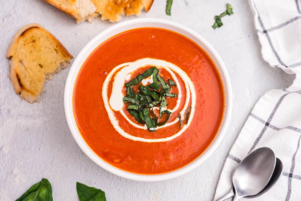 A bowl of creamy tomato soup garnished with a swirl of cream and fresh basil, served with toasted bread and a spoon on a light textured surface.