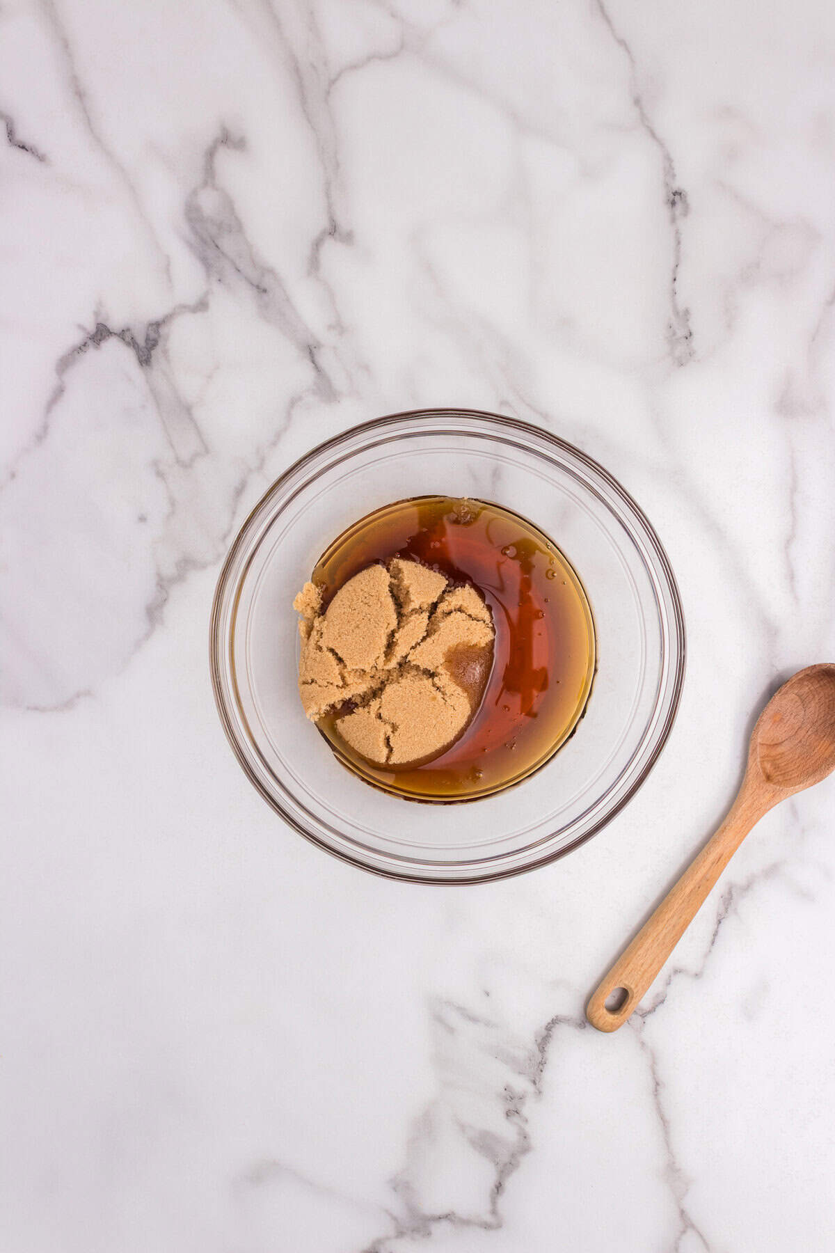 A glass bowl with packed brown sugar and maple syrup sitting on a marble countertop next to a wooden spoon.