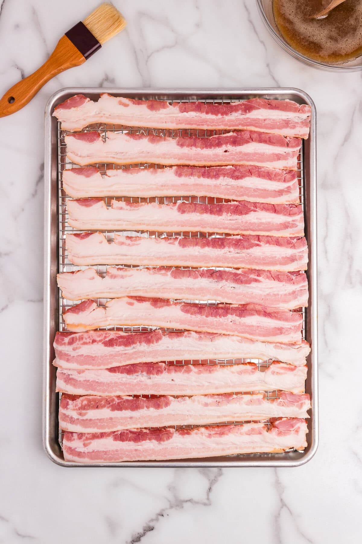 Raw bacon strips neatly arranged on a wire rack over a baking sheet, with a basting brush and sugar mixture nearby.