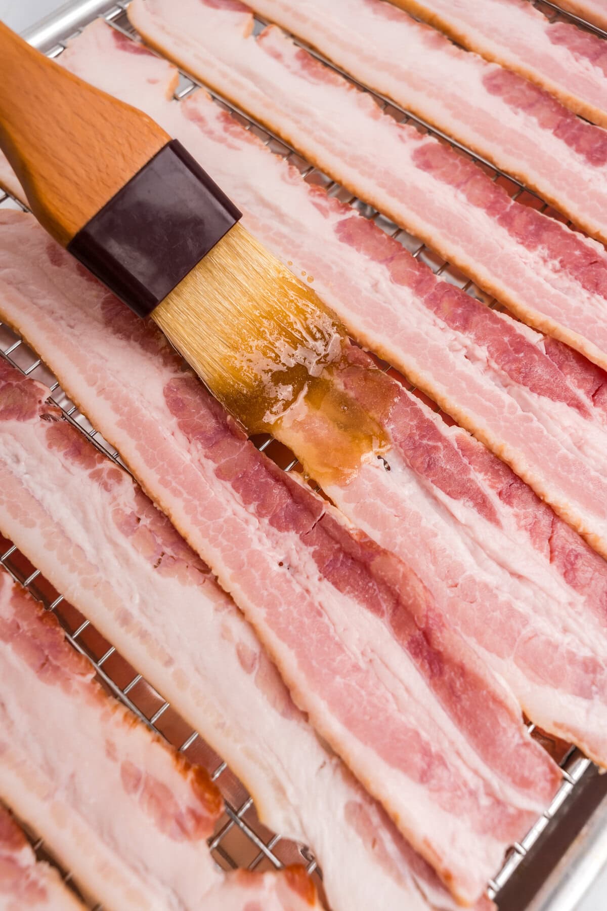 Close-up of a basting brush applying the sugar mixture to raw bacon strips on a rack.