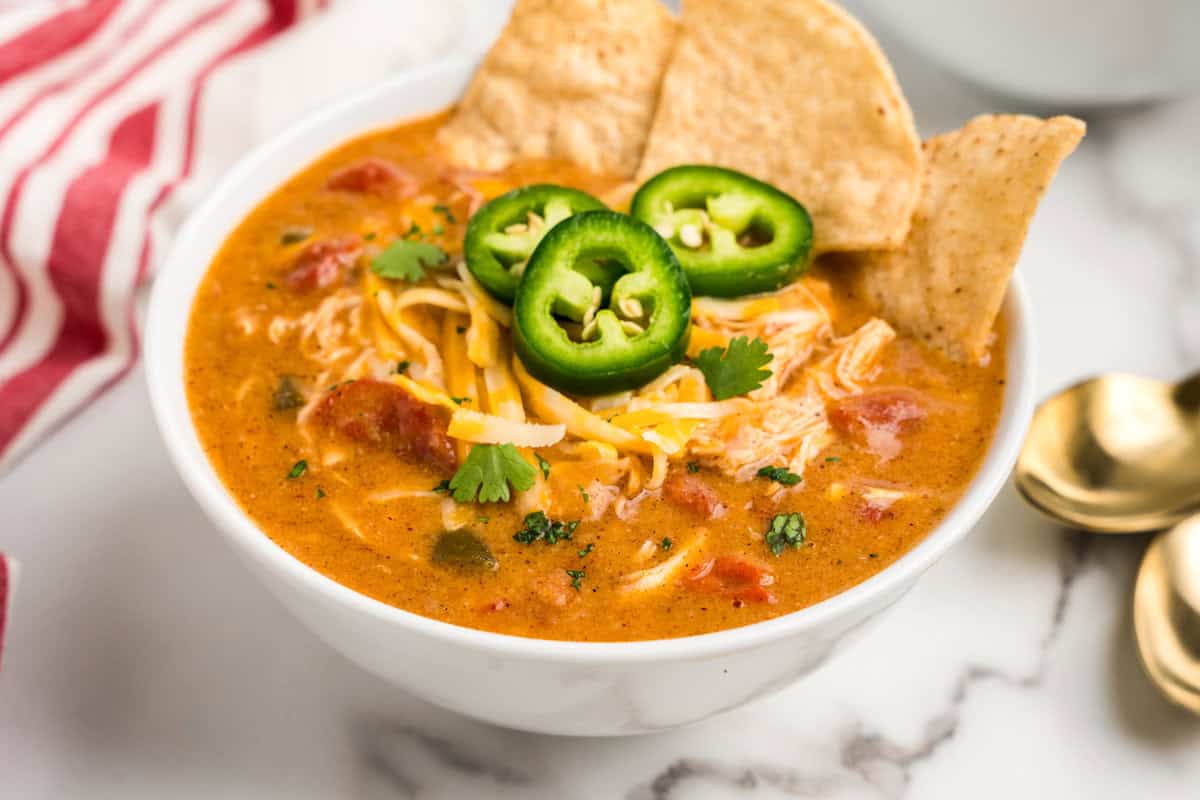 A bowl of Crock Pot King Ranch Chicken Soup with a rich, creamy broth, shredded chicken, diced tomatoes, and spices. The soup is garnished with shredded cheese, sliced jalapeños, fresh cilantro, and crispy tortilla chips. A red and white striped napkin and gold spoons are placed nearby on a marble countertop.