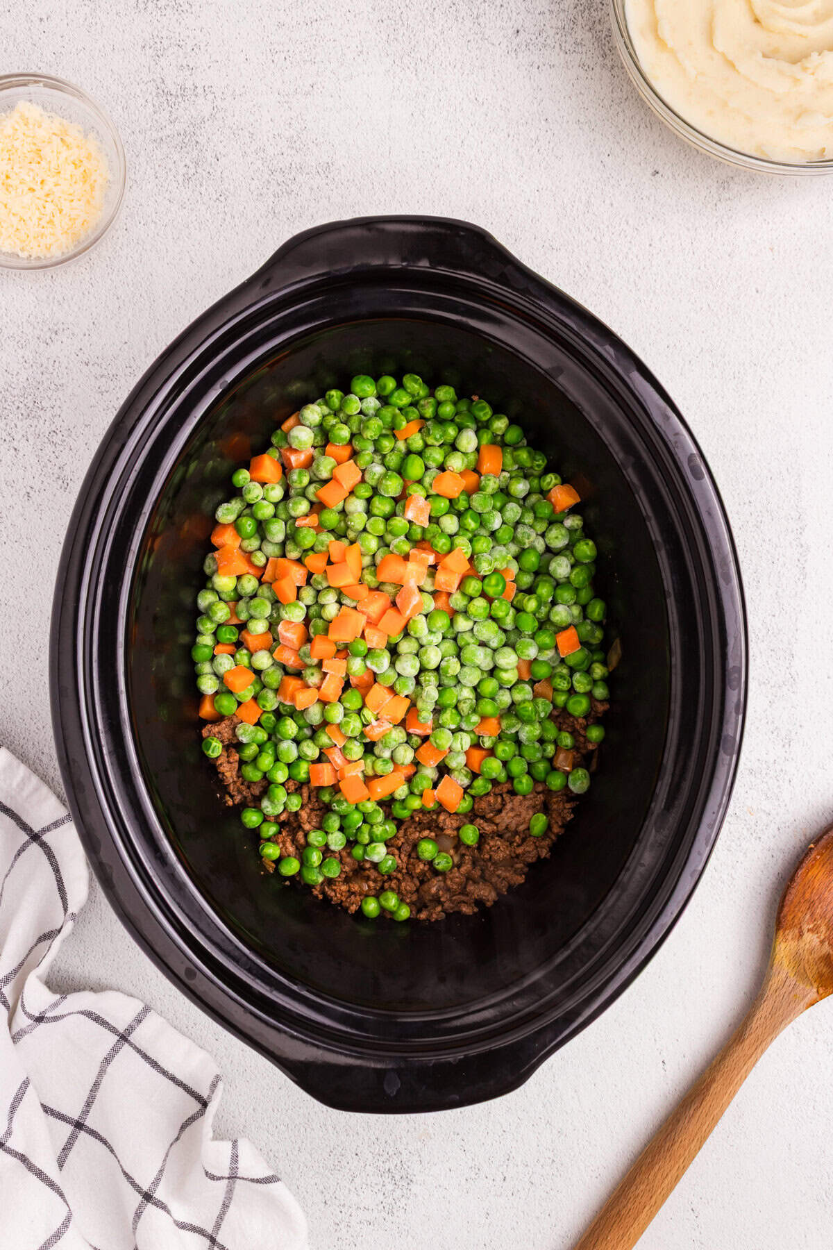 Layer of cooked ground beef mixture topped with frozen peas and carrots in a black slow cooker.
