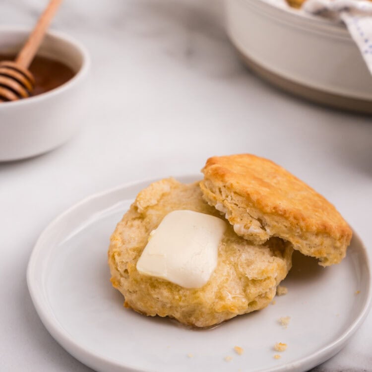 A biscuit is split open on a plate with a pat of butter melting into the warm interior; honey in a bowl with a dipper is in the background.