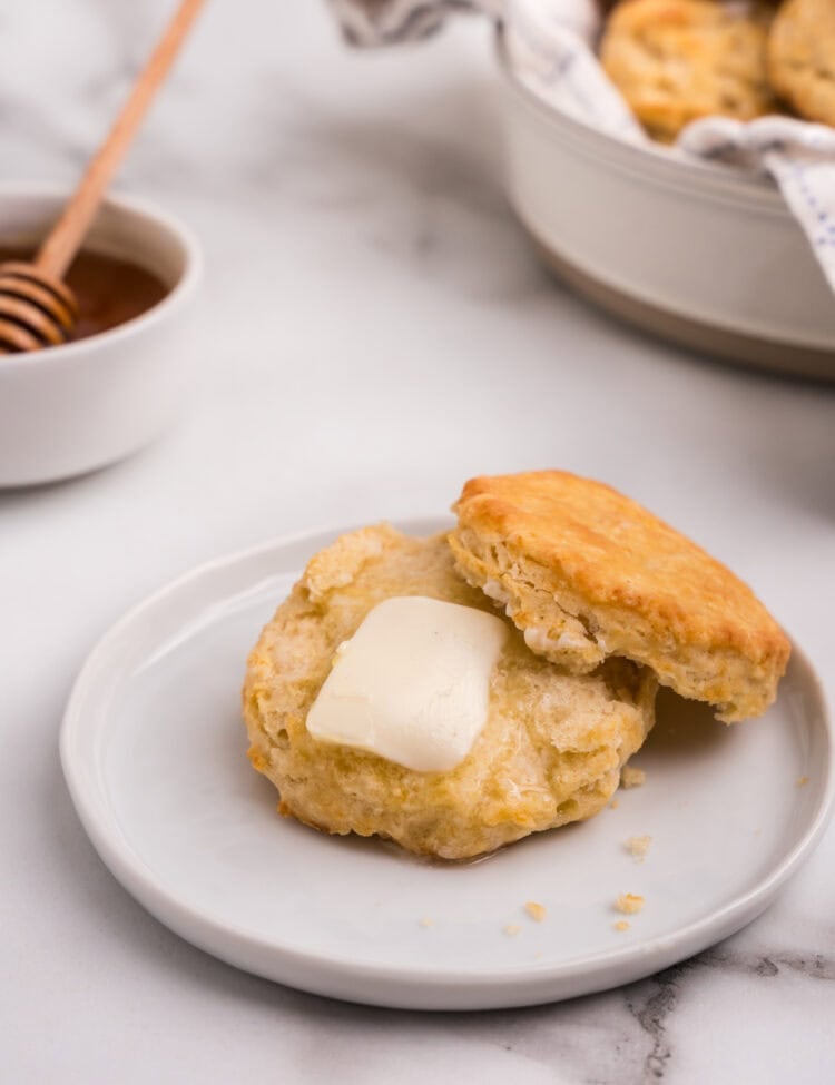 A biscuit is split open on a plate with a pat of butter melting into the warm interior; honey in a bowl with a dipper is in the background.