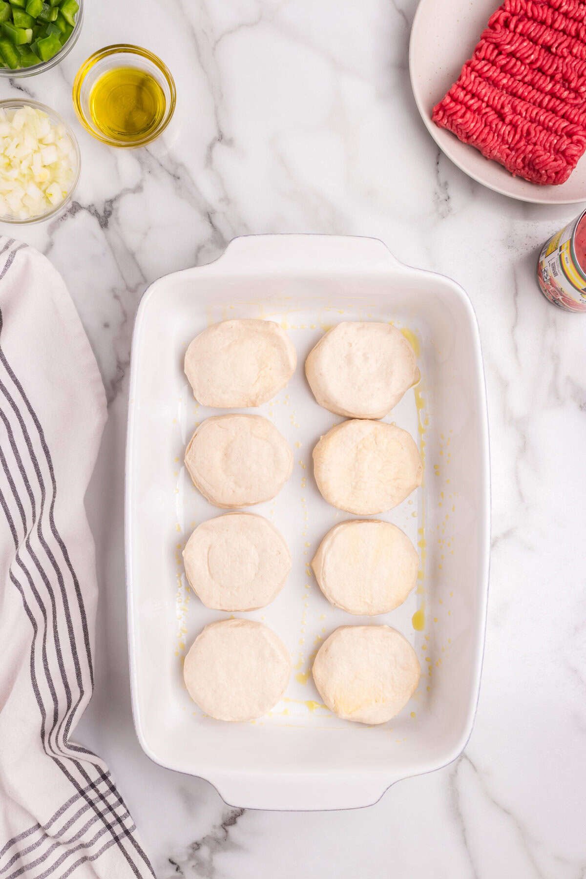 A white baking dish containing raw biscuit dough rounds, neatly arranged in two rows, with ingredients like chopped green peppers, onions, and ground beef around it.