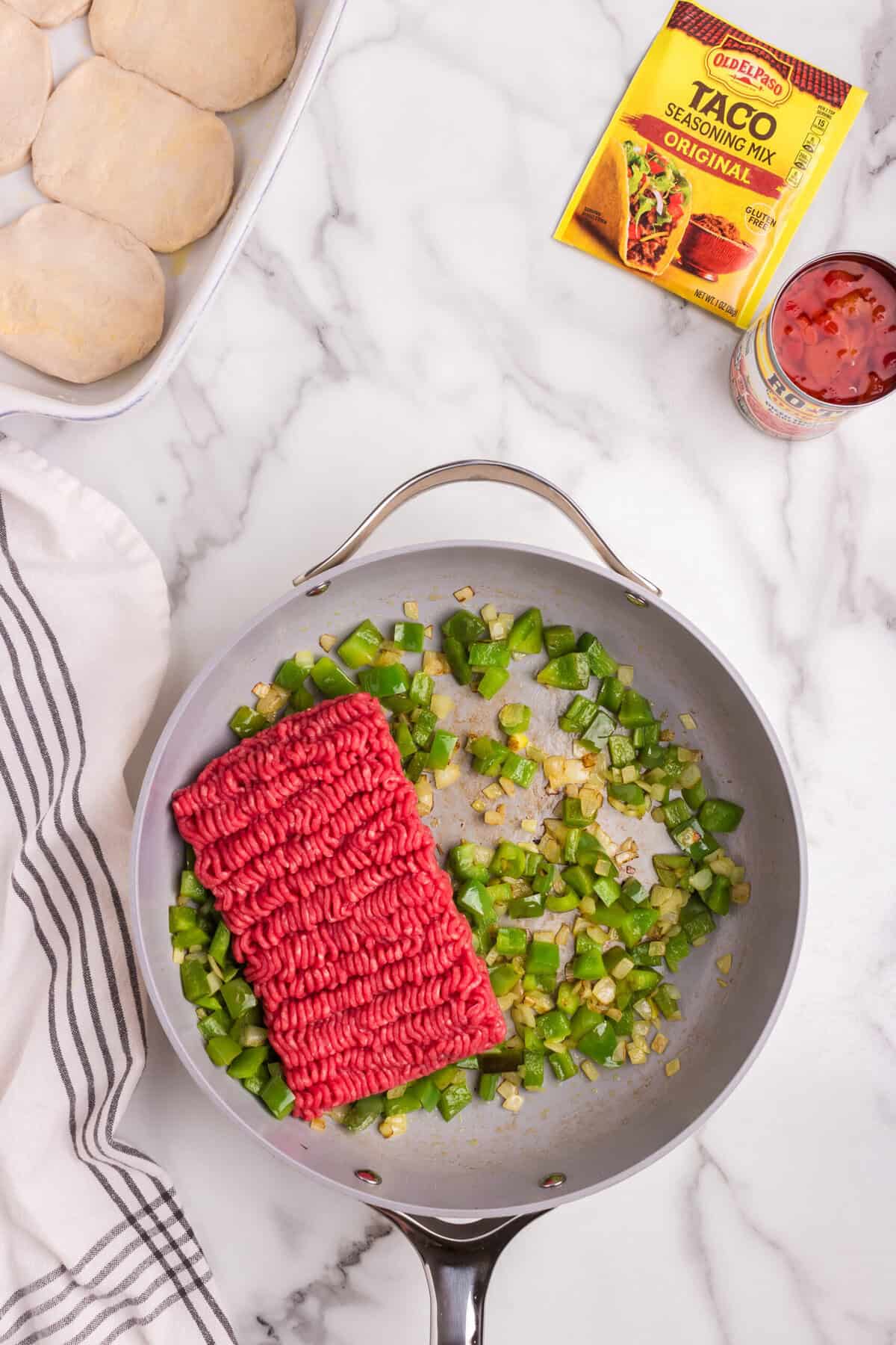 A skillet with raw ground beef and diced green bell peppers, with a packet of taco seasoning and a can of diced tomatoes nearby.