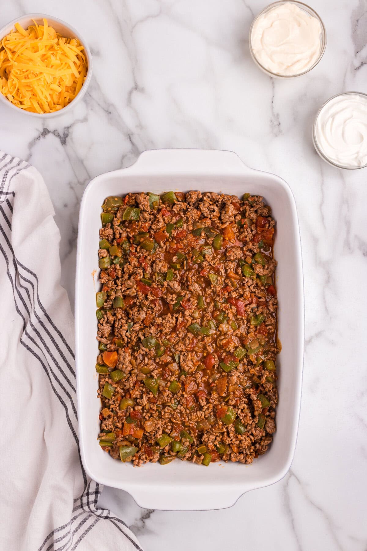 The prepared meat mixture spread evenly over the biscuit layer in the baking dish, with shredded cheese and sour cream nearby.