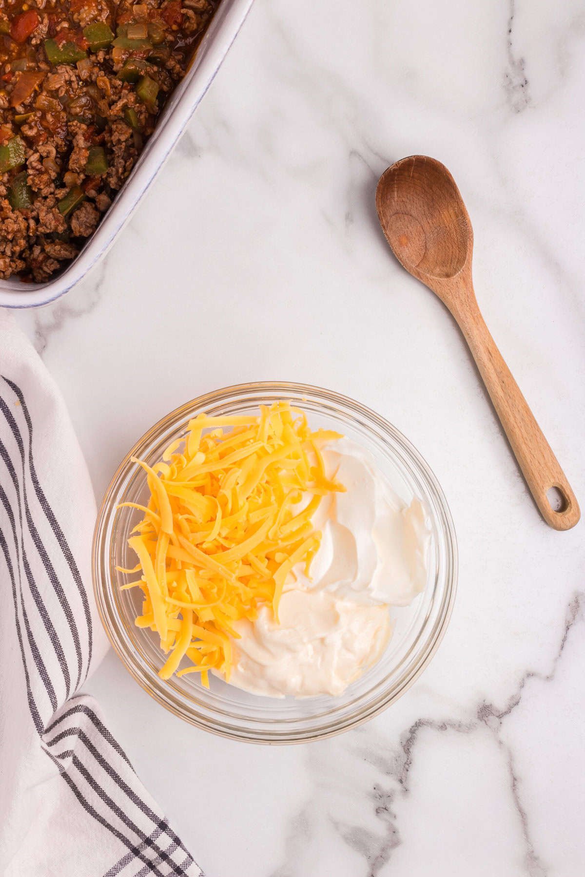 A small glass bowl with a mixture of shredded cheese and sour cream, with a wooden spoon next to it, ready to be used as a topping.