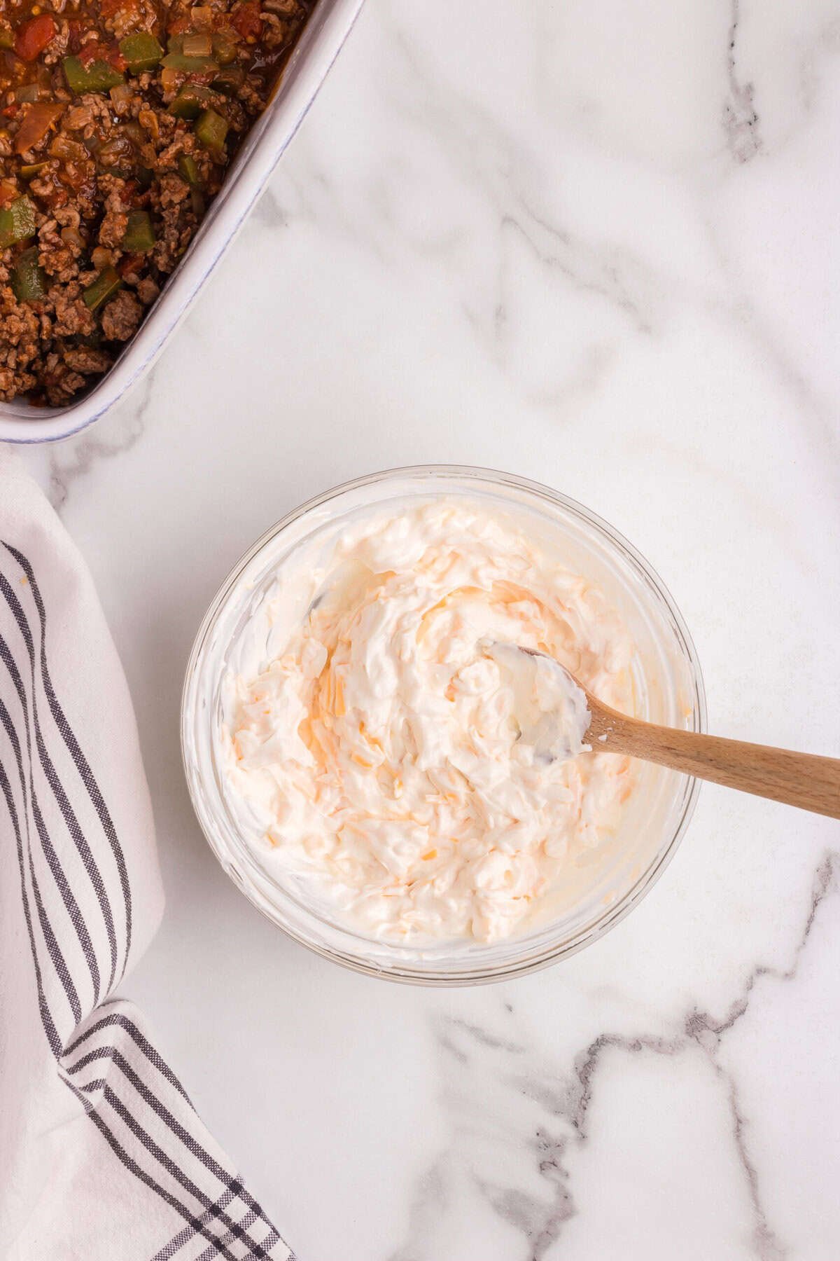 A glass bowl filled with a creamy mixture of sour cream and shredded cheese, with a wooden spoon stirring it. In the background, a portion of a baking dish with seasoned ground beef and bell peppers is visible on a marble countertop.