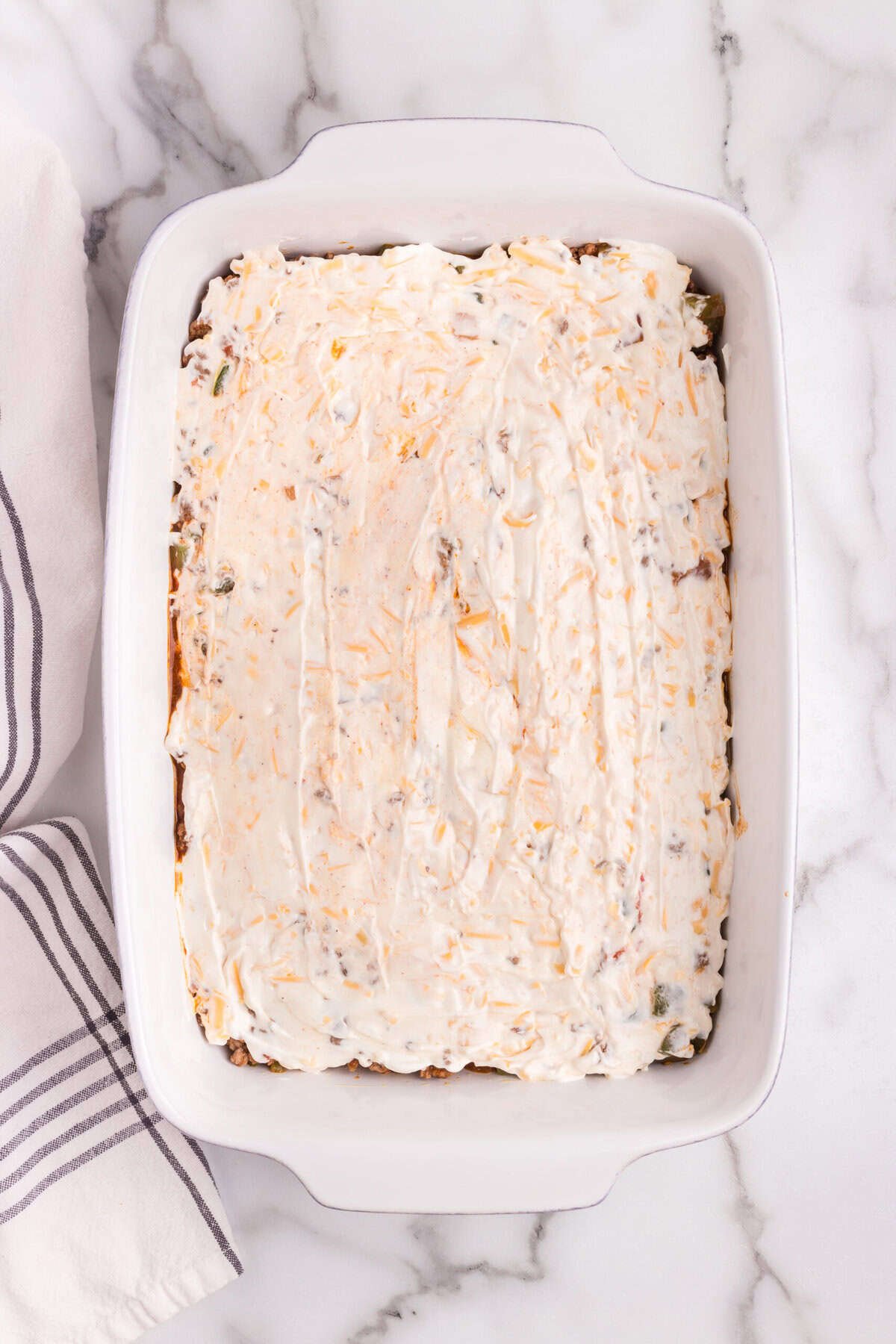 A white rectangular baking dish filled with a casserole, topped with a creamy mixture of sour cream and shredded cheese, spread evenly over the surface.